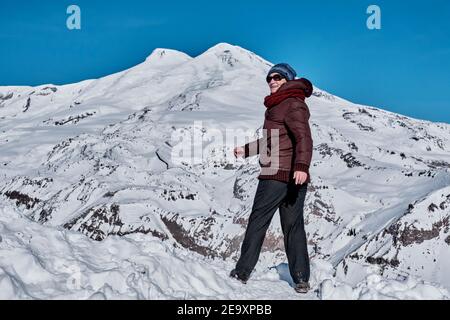Eine ältere Wandererin in Sonnenbrillen, lässige Winterkleidung steht eine halbe Umdrehung auf dem Schnee vor dem Hintergrund des Mount Elbrus. Nordkaukasus, Kaba Stockfoto