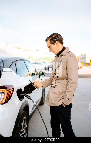 Seitenansicht der stilvollen männlichen Unternehmer Browsing Smartphone im Stehen An der Tankstelle und tanken Auto Stockfoto