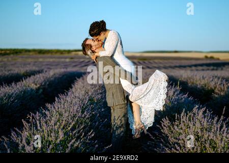 Seitenansicht des Bräutigams, der die Braut hebt, während er in Lavendel steht Feld auf dem Hintergrund des klaren blauen Himmels am Hochzeitstag Stockfoto