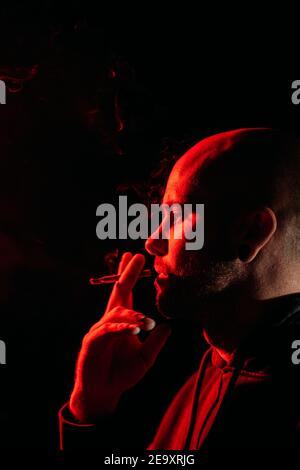 Seitenansicht des männlichen Rockers mit Glatze Rauchen und Ausatemrauch im dunklen Studio mit rotem Neonlicht an Schwarzer Hintergrund Stockfoto
