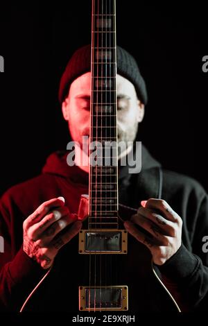 Beschaulicher männlicher Rockmusiker, der mit E-Gitarre auf Schwarz steht Hintergrund im Studio mit rotem Licht Stockfoto