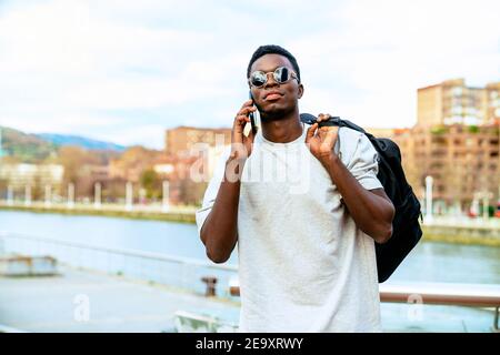 Junger afroamerikanischer Rüde mit Rucksack in trendigen Sonnenbrillen im Gespräch Auf dem Handy gegen Stadtfluss unter bewölktem Himmel Stockfoto