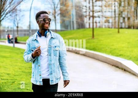 Junger afroamerikanischer Mann in Denim-Jacke mit Kopfhörern Auf dem Weg zwischen den Rasenflächen in der Stadt Stockfoto