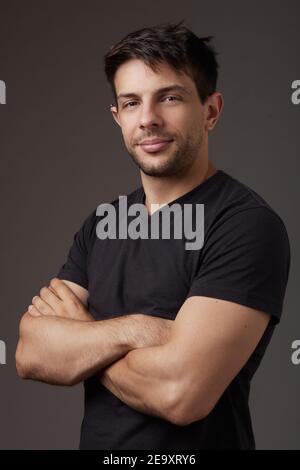 Mann im schwarzen Hemd mit gekreuzten Armen, der im Studio auf grauem Hintergrund auf die Kamera schaut Stockfoto