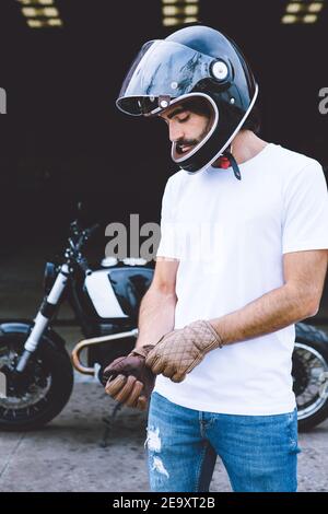 Konzentrierter junger unrasiert ethnischer männlicher Biker in lässigem Outfit und Helm trägt Handschuhe, während er in der Nähe des Motorrads auf der Straße steht Stockfoto