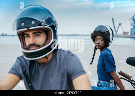 Stilvolle junge multiethnische Paar in lässigem Outfit und Helme ruhen Auf der Promenade in der Nähe von Meer nach dem Motorradfahren Stockfoto