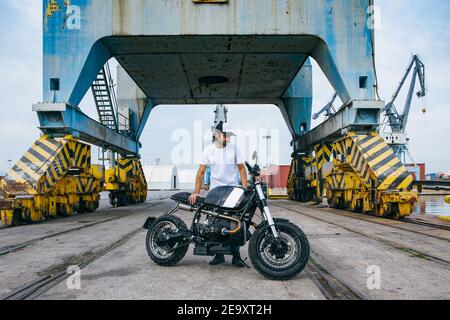 Ganzer Körper von schweren jungen ethnischen männlichen Biker in casual Kleidung und Kappe stehen in der Nähe von Motorrad in der Nähe von Industriekranen Am Meer Stockfoto