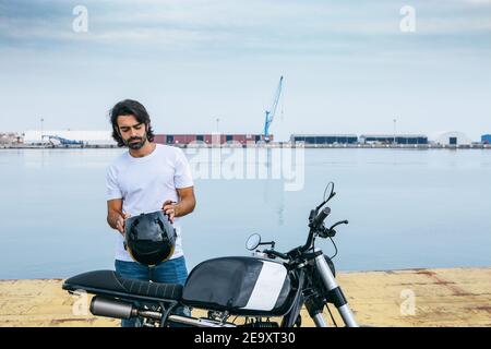 Zuversichtlich junge bärtige ethnische Kerl in weißen T-Shirt und Jeans im Stehen Motorrad am Meer Stockfoto
