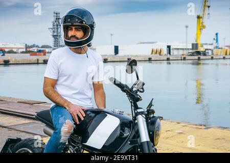 Zuversichtlich junge bärtige ethnische Kerl in weißen T-Shirt und Jeans im Stehen Motorrad am Meer Stockfoto