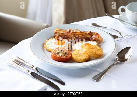 Ein großer Winkel appetitlich gebratener Eier mit Speck Scheiben serviert Auf Teller mit gefüllten Tomaten und knusprigen Mozzarella und platziert Auf dem Tisch mit Cutler und Cu Stockfoto