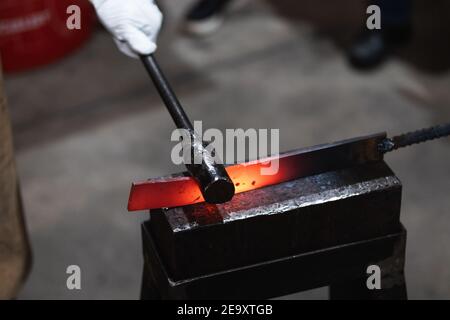 Unerkennbarer Handwerker hämmert während des Schmiedens heißes Eisenstück auf Amboss Prozess in der Werkstatt Stockfoto