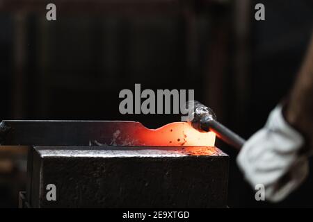 Unerkennbarer Handwerker hämmert während des Schmiedens heißes Eisenstück auf Amboss Prozess in der Werkstatt Stockfoto