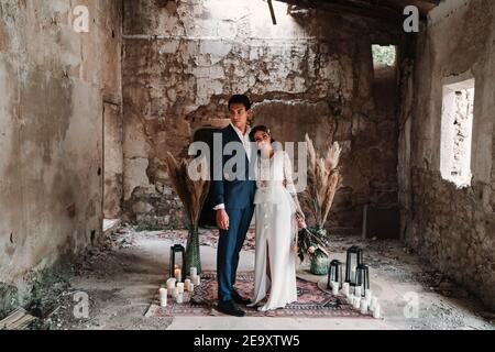 Seitenansicht Ganzkörper liebevolle junge ethnische Neuvermählte Paar tragen Chic Hochzeitskleidung umarmt und küsst sanft in obsolete Rest Gebäude Stockfoto