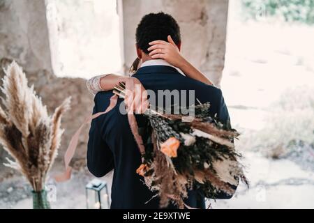 Rückansicht Bräutigam in eleganten Smoking umarmt Braut mit Blumenstrauß Im Grunge Außenbau an sonnigen Tagen Stockfoto