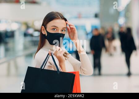 Positive junge Erwachsene Frau trägt schützende medizinische Gesichtsmaske mit Papier Einkaufstaschen in den Händen im Einkaufszentrum. Black friday und covid-19 Stockfoto