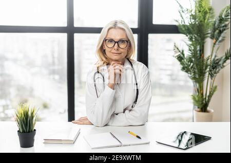 Kopfschuss von zuversichtlich mittleren Alters MD, pädiatrischen, Arzt trägt weiße medizinische Kleid und Brillen mit einem Stethoskop auf den Schultern sitzen an der Stockfoto