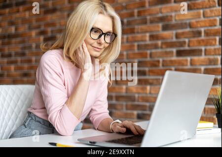 Bezaubernde Geschäftsfrau mittleren Alters trägt stilvolle Brillen und elegante Freizeitkleidung mit einem Laptop für die Büroarbeit, weibliche Angestellte prüft Post Stockfoto