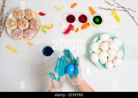 Der Prozess der Färbung von Eiern für Ostern in blauen und roten Farben. Eine Frau in Handschuhen malt Eier mit Farbstoff aus dem Glas, Draufsicht, Weidenzweige und Farbe Stockfoto