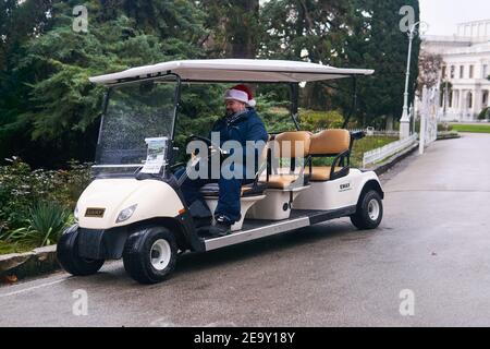 Jalta, Russland - 26. Januar 2021: Fahrer eines touristischen elektrischen Golfwagens in einem Weihnachtshut schaut in ein Smartphone und lächelt in seiner Freizeit in der Stockfoto