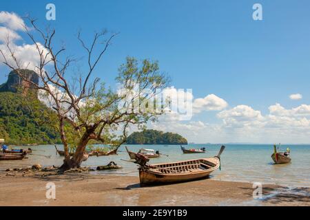 Eine riesige Felsformation überschattet den weißen Sand und das blaue Meer sowie die traditionellen Long Tail Boote am Railay Beach in Krabi, Thailand Stockfoto