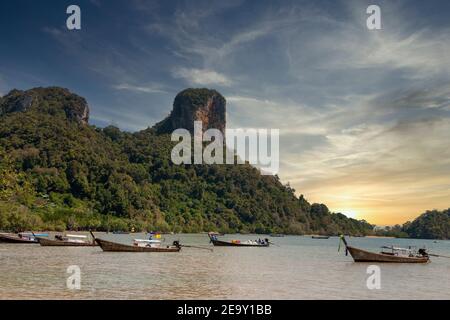 Eine riesige Felsformation überschattet den weißen Sand und das blaue Meer sowie die traditionellen Long Tail Boote am Railay Beach in Krabi, Thailand Stockfoto