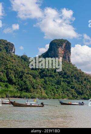 Eine riesige Felsformation überschattet den weißen Sand und das blaue Meer sowie die traditionellen Long Tail Boote am Railay Beach in Krabi, Thailand Stockfoto