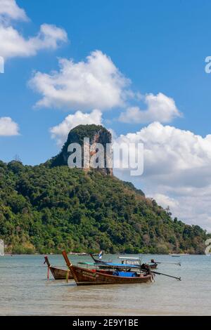 Eine riesige Felsformation überschattet den weißen Sand und das blaue Meer sowie die traditionellen Long Tail Boote am Railay Beach in Krabi, Thailand Stockfoto