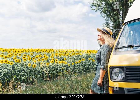 Junge blonde Frau, die mit dem Wohnmobil reist. Blick auf Sonnenblumenfeld an sonnigen Sommertag. Stockfoto