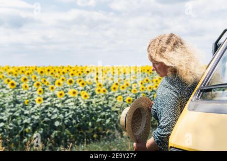 Junge blonde Frau, die mit dem Wohnmobil reist. Blick auf Sonnenblumenfeld an sonnigen Sommertag. Stockfoto