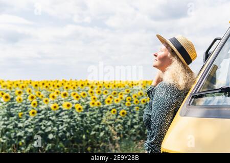 Junge blonde Frau, die mit dem Wohnmobil reist. Blick auf Sonnenblumenfeld an sonnigen Sommertag. Stockfoto