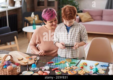 Kleiner Junge macht Foto am Telefon seines Handwerks Er schmückt Lebkuchen zusammen mit seiner Mutter Stockfoto