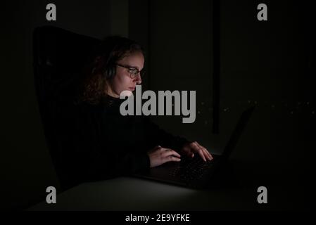 Frau mit Kopfhörern, die im Dunkeln der Nacht auf ihrem Laptop tippt. Überarbeitungskonzept. Stockfoto