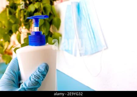 Eine Hand in einem medizinischen Handschuh hält eine Flasche mit einem Antiseptikum auf einem Hintergrund von grünen Blättern und einem weißen Hintergrund mit Masken, Hygieneprodukt, protec Stockfoto