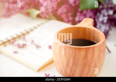 Finnische Holztasse mit Kaffee auf dem Tisch. In der Nähe liegt ein geschlossenes Notizbuch und ein Strauß Flieder. Guten Morgen und gute Laune, Planung a d Stockfoto