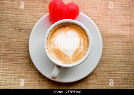 Eine weiße Tasse auf einer Untertasse mit Kaffee und Marmelade Herz steht auf einer Fensterbank auf Sackleinen Stoff, Einsamkeit. Trinken mit Milch, mit Schaum Herz, oben Stockfoto