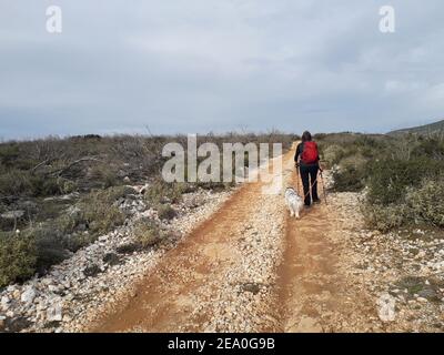 Frau Wanderer mit rotem Rucksack und ihr Hund auf einer Schotterstraße auf Mt Hymettus, n Griechenland Stockfoto