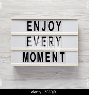 „Enjoy Every Moment“ auf einem Leuchtkasten auf einer weißen Holzoberfläche, Draufsicht. Flachlage, über Kopf, von oben. Stockfoto