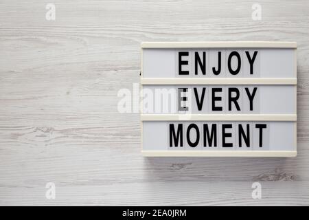 „Enjoy Every Moment“ auf einem Leuchtkasten auf weißem Holzhintergrund, Draufsicht. Flachlage, über Kopf, von oben. Speicherplatz kopieren. Stockfoto