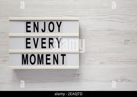 „Enjoy Every Moment“ auf einem Leuchtkasten auf weißem Holzhintergrund, Draufsicht. Flachlage, über Kopf, von oben. Speicherplatz kopieren. Stockfoto