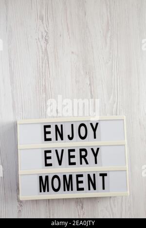 „Enjoy Every Moment“ auf einem Leuchtkasten auf weißem Holzhintergrund, Draufsicht. Flachlage, über Kopf, von oben. Speicherplatz kopieren. Stockfoto