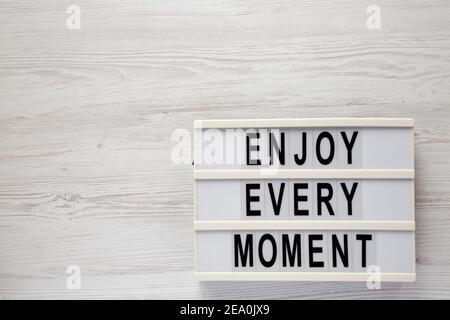 „Enjoy Every Moment“ auf einem Leuchtkasten auf einer weißen Holzoberfläche, Draufsicht. Flachlage, über Kopf, von oben. Leerzeichen für Text. Stockfoto