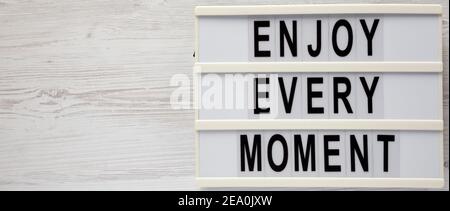 „Enjoy Every Moment“ auf einem Leuchtkasten auf weißem Holzhintergrund, Draufsicht. Flachlage, über Kopf, von oben. Speicherplatz kopieren. Stockfoto