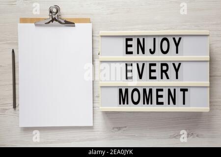 "Genießen Sie jeden Moment" auf einem Leuchtkasten, Zwischenablage mit leerem Blatt Papier auf einer weißen Holzfläche, Draufsicht. Flachlage, über Kopf, von oben. Stockfoto