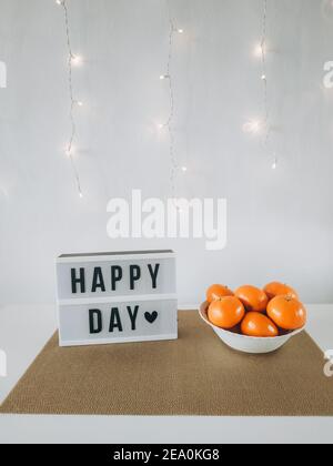 HAPPY DAY Wort auf Leuchtkasten mit Mandarinen an der Wand mit Licht Hintergrund. Gemütliches Haus compozition. Stockfoto