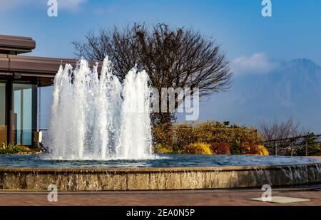 Ein Brunnen spritzt Wasser an die frische Luft, vor dem Hintergrund eines blattlosen Baumes, und der Vulkan Sakurajima, in Kagoshima Stadt, Japan. Stockfoto