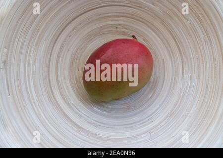 Eine einzige ganze Mangofrucht von roten und grünen Farben In einer Bambusschale helles Holz organisch Hintergrund Draufsicht Mit Kopierbereich Stockfoto