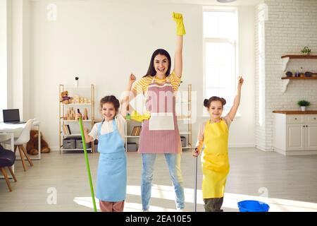 Mutter mit zwei Töchtern, die das Haus putzen, stehen mit erhobenen Armen und geben vor, Superhelden zu sein. Stockfoto