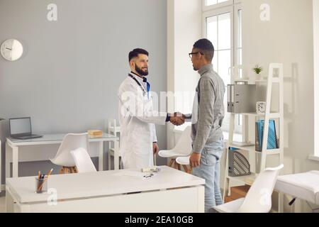 Der Arzt schüttelt die Hände mit seinem dunkelhäutigen Patienten und begrüßt ihn in einer modernen medizinischen Klinik. Stockfoto