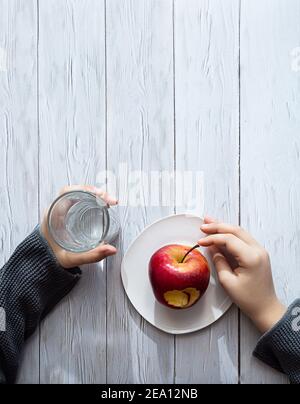 Minimaler gesunder Snack-Konzept. Die Hände des Kindes halten einen Apfel und ein Glas Wasser. Stillleben auf einem hellen Holztisch mit Schatten und Highli Stockfoto