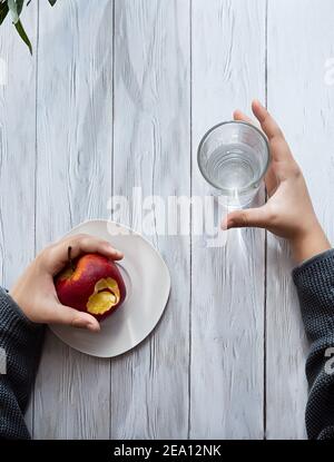 Minimaler gesunder Snack-Konzept. Die Hände des Kindes halten einen Apfel und ein Glas Wasser. Stillleben auf einem hellen Holztisch mit Schatten und Highli Stockfoto